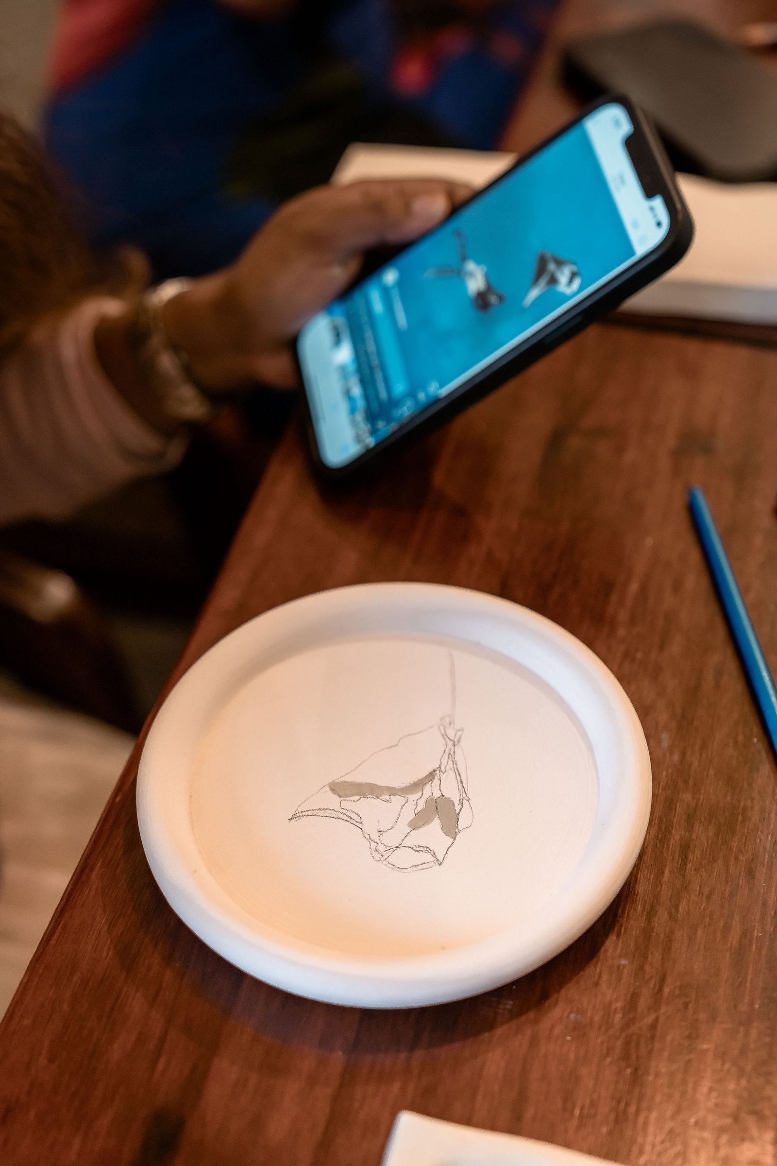 A reference photograph on a phone being compared against an unfired clay plate in the workshop