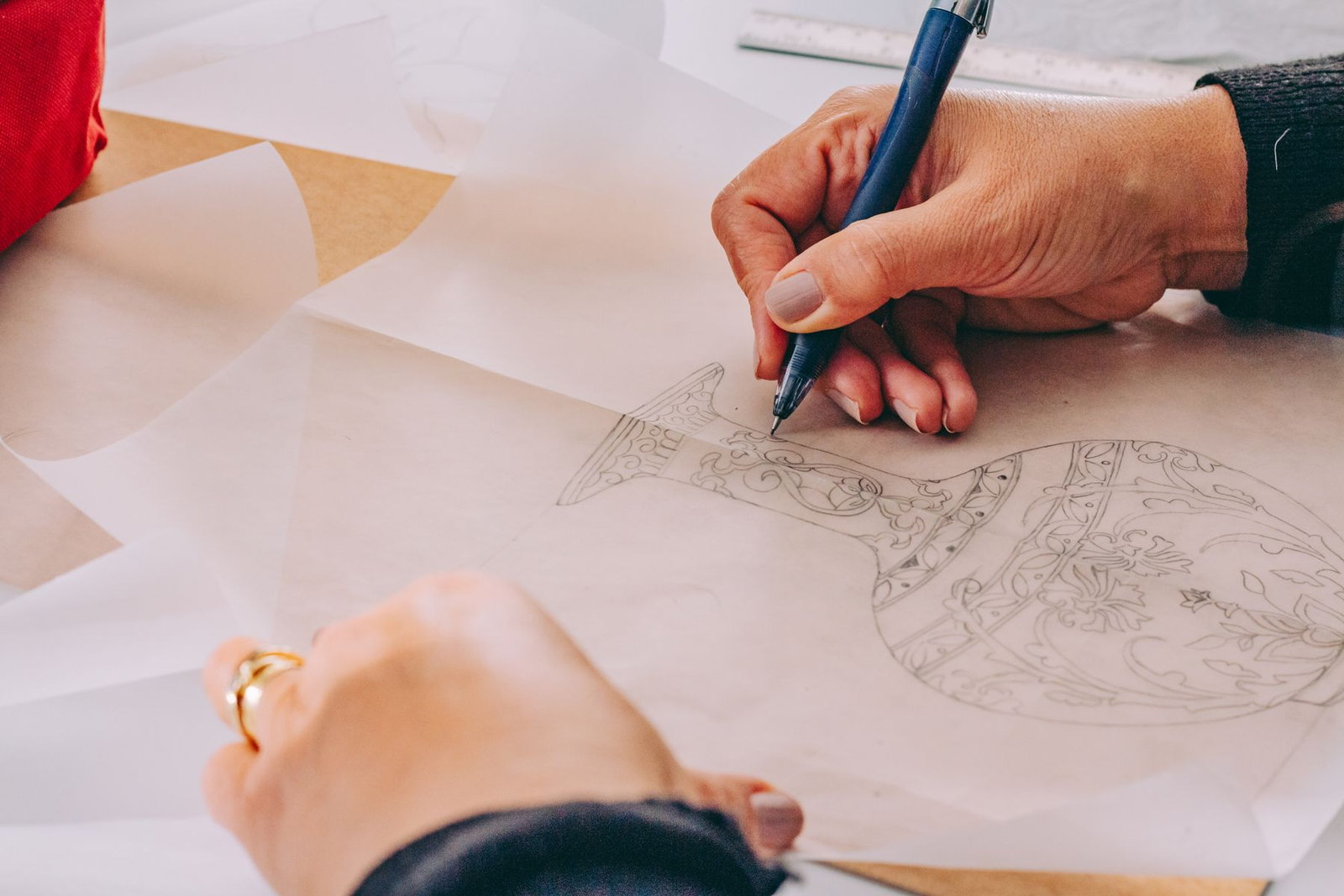 Hands drawing a floral pattern onto a flat clay surface with a pencil and a brush